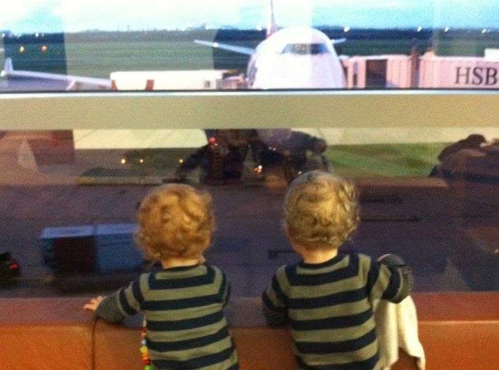 Two toddlers stand on terminal seating at the window glass, watching a widebody jet parked at the gate